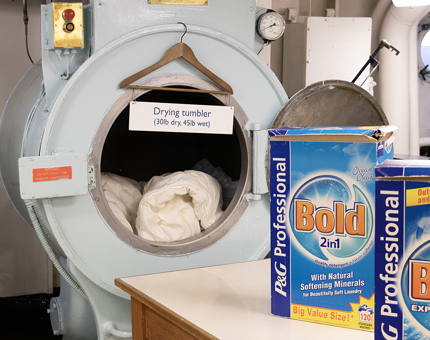 A large drying tumbler machine in the onboard Laundry. Two boxes of laundry detergent are on a bench next to the machine.
