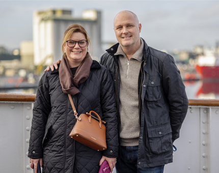 A man and woman pose for a photo on the Bridge deck of Britannia. There is a tall white building in the background.