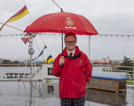 A smiling Visitor Assistant stands on Britannia's Verandah Deck holding a red umbrella in the rain. Port of Leith Edinburgh.