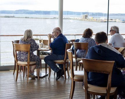 Guests sitting at tables in the Royal Deck Tearoom. Thorough the big windows you can see the water.