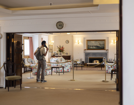 A man is listening to the audio guide handset in the State Drawing Room. There is a clock above the door, and in the room there are floral couches, side tables and a grey fireplace.