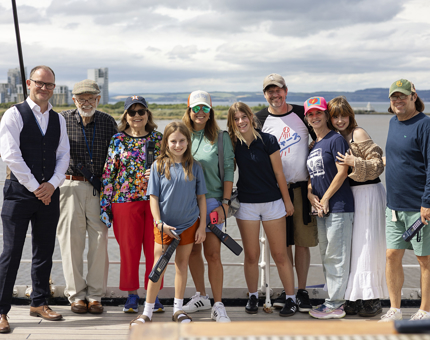 10 visitors of various ages posing for a photo together on Britannia's Bridge. In the background there is a view of the water.