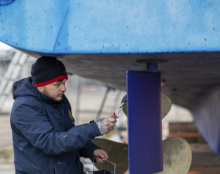 A maintenance team member standing under a boat holding a paintbrush and tin of blue paint.