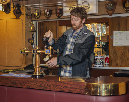 A man stands behind the bar in the Crew's mess pretending to pour a pint.