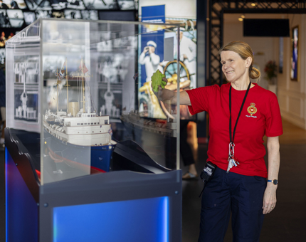 A Housekeeper is holding a cloth, wiping a glass case which holds a replica model of Britannia.